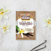 Un sachet de sucre vanillé Vahiné Sucre Vanillé est présenté avec des gousses de vanille et des fleurs.