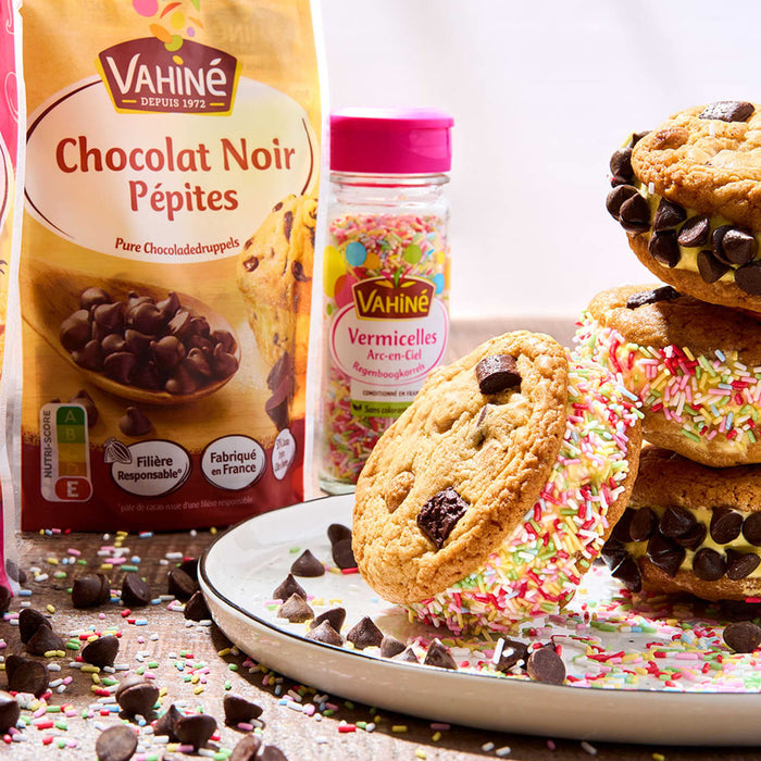 Une pile de sandwichs glacés aux biscuits aux pépites de chocolat faits maison avec des vermicelles, à côté d'un sac de pépites de chocolat noir Vahiné et d'un récipient de vermicelles.