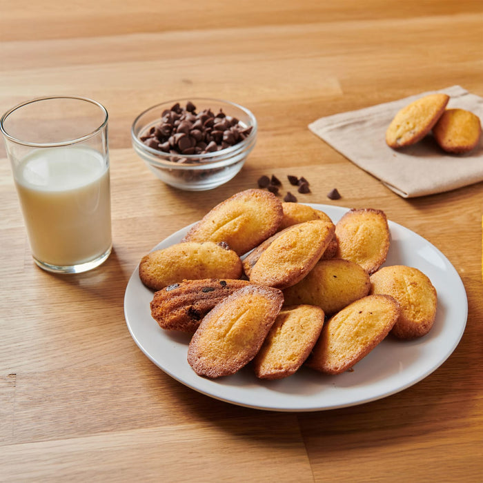 Une assiette de madeleines fraARchement cuites est prACsentACe A? cA?tAC d'un verre de lait et d'un bol de pACpites de chocolat, suggACrant une dAClicieuse collation ou un dessert.