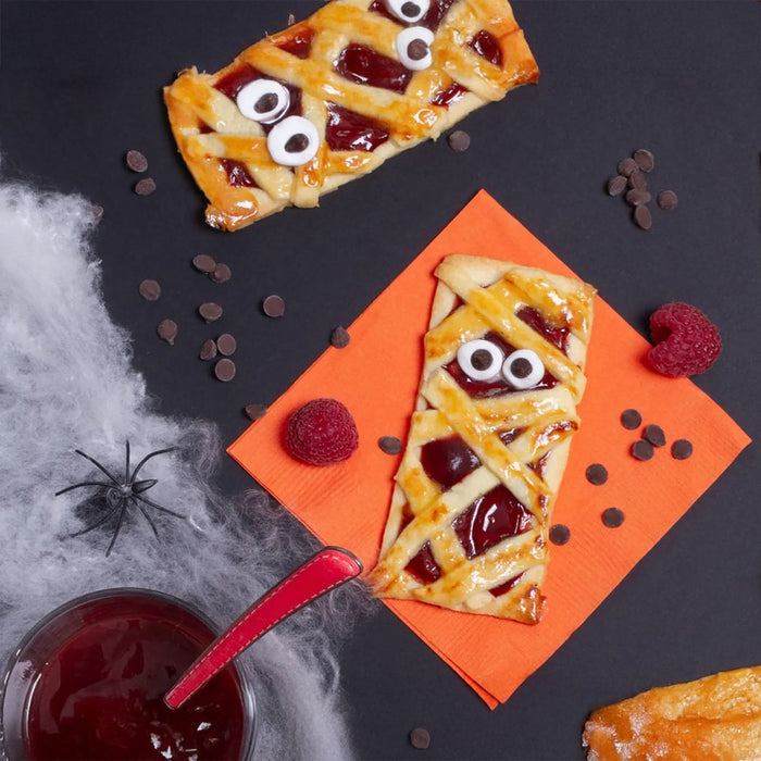 Une image sur le thème d'Halloween présentant des pâtisseries de style momie remplies de confiture rouge et décorées d'yeux en bonbon, entourées de décorations d'Halloween comme des toiles d'araignée et des pépites de chocolat.