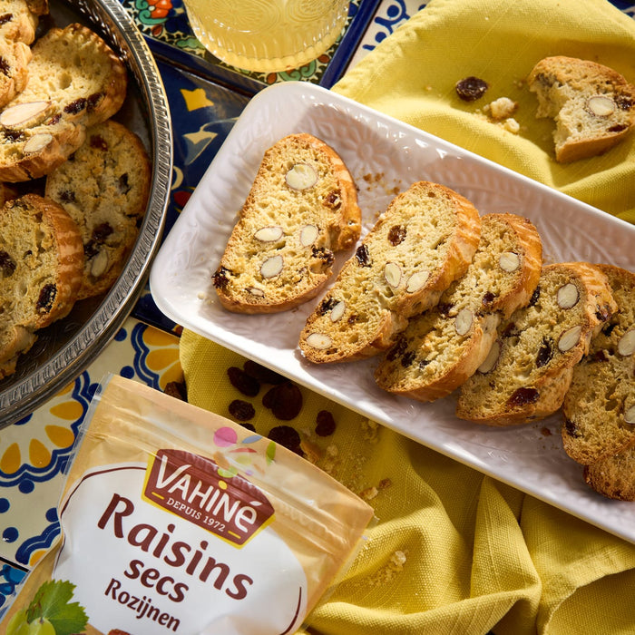 Une assiette de biscotti avec des Vahiné Raisins secs est présentée sur un tissu jaune, à côté d'un sachet de raisins.