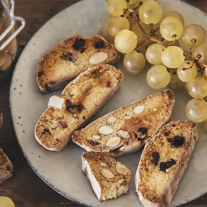 Une assiette de biscotti aux amandes et aux fruits secs est servie avec une grappe de raisins verts.