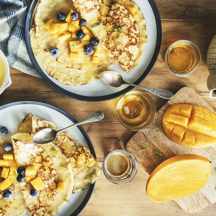 Deux assiettes de crAapes garnies de mangue et de myrtilles sont prACsentACes sur une table en bois, accompagnACes de tranches de mangue et de petits pots de miel.