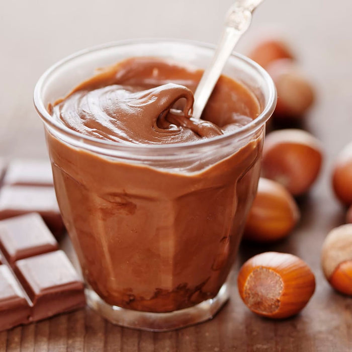 Un pot en verre rempli de pâte à tartiner chocolat-noisette faite maison, entouré de noisettes et de carrés de chocolat, présentant une gourmandise délicieuse et indulgente.