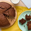 Une photo en plongée montre un gâteau aux pépites de chocolat fraîchement cuit, préparé avec des ingrédients Vahine, tranché sur une planche en bois à côté d'une assiette avec des morceaux de gâteau et d'un verre de lait.