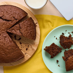 Une photo en plongée montre un gâteau aux pépites de chocolat fraîchement cuit, préparé avec des ingrédients Vahine, tranché sur une planche en bois à côté d'une assiette avec des morceaux de gâteau et d'un verre de lait.