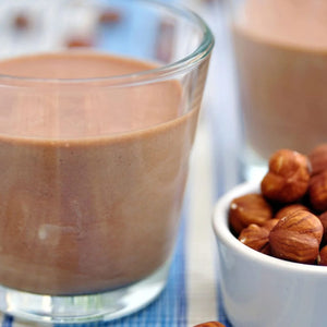 Un gros plan d'un verre de lait de noisette onctueux à côté d'un petit bol blanc de noisettes, représentant les Vahine Nutritional Facts Ingredients And Hero Reipes Visuals For Newly Launched Skus.