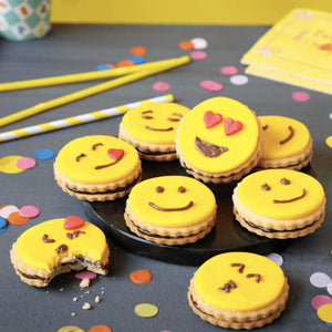 Une collection de biscuits sur le thA?me des emojis, avec un glaA?age jaune et une garniture au chocolat, sont disposACs sur une assiette, entourACs de confettis colorACs et de pailles, suggACrant une cAClACbration festive.