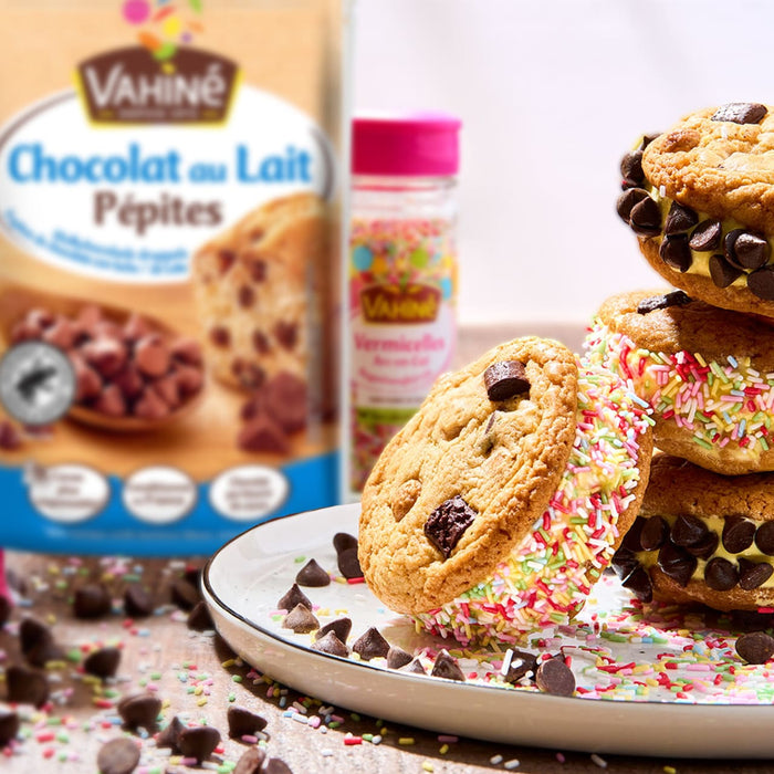 Une assiette de sandwichs aux biscuits pépites de chocolat, certains roulés dans des vermicelles colorés et d'autres avec des pépites de chocolat supplémentaires, est présentée avec un paquet de Vahine Sch Pepites Chocolaitx10-Va et une bouteille de vermicelles Vahiné Arc-en-Ciel en arrière-plan.