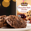 Un paquet de Vahine Sch100G Pepites Chunkx10-Va, contenant des pépites de chocolat noir, est présenté derrière une assiette de biscuits au chocolat fraîchement préparés.