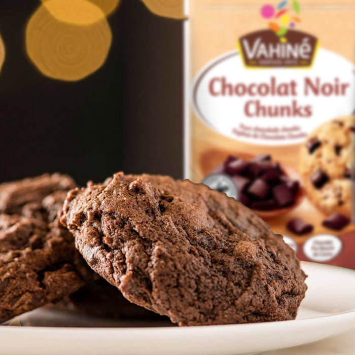 Un paquet de Vahine Sch100G Pepites Chunkx10-Va, contenant des pépites de chocolat noir, est présenté derrière une assiette de biscuits au chocolat fraîchement préparés.