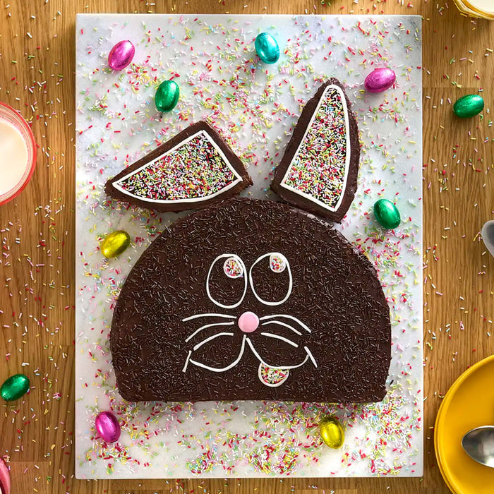 Un gâteau de Pâques fantaisiste en forme de lapin en chocolat, décoré de vermicelles et d'œufs en bonbon, repose sur une surface recouverte de papier sulfurisé.