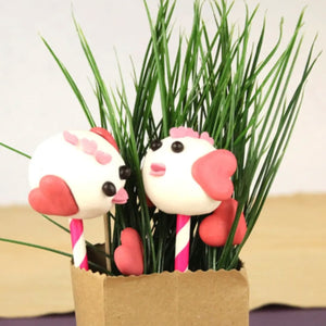 Deux cake pops en forme d'oiseau décorés de cœurs roses et d'yeux noirs sont présentés dans un sac en papier brun rempli d'herbe artificielle.