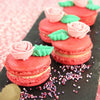 Trois macarons rouges dACcorACs de roses roses et de feuilles vertes reposent sur une surface d'ardoise parsemACe de confettis colorACs.