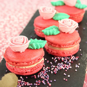 Trois macarons rouges dACcorACs de roses roses et de feuilles vertes reposent sur une surface d'ardoise parsemACe de confettis colorACs.
