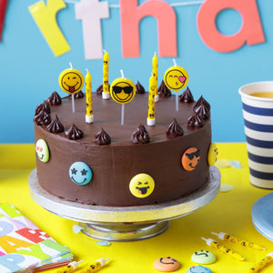 Un gâteau d'anniversaire au chocolat décoré de bougies emoji et de décorations emoji comestibles repose sur un présentoir à gâteau, prêt pour une célébration.