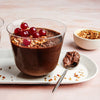 Un dessert mousse au chocolat décadent est présenté dans un verre, garni de groseilles rouges et de noix hachées, accompagné d'une cuillère sur une assiette blanche.
