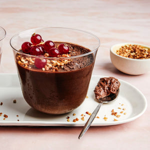 Un dessert mousse au chocolat décadent est présenté dans un verre, garni de groseilles rouges et de noix hachées, accompagné d'une cuillère sur une assiette blanche.