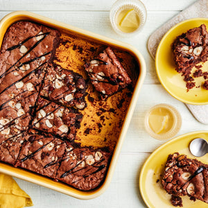 Un plat de brownies fraîchement cuits avec des noix et un filet de chocolat est présenté à côté de deux assiettes contenant des portions de brownies et des verres d'une boisson de couleur claire.