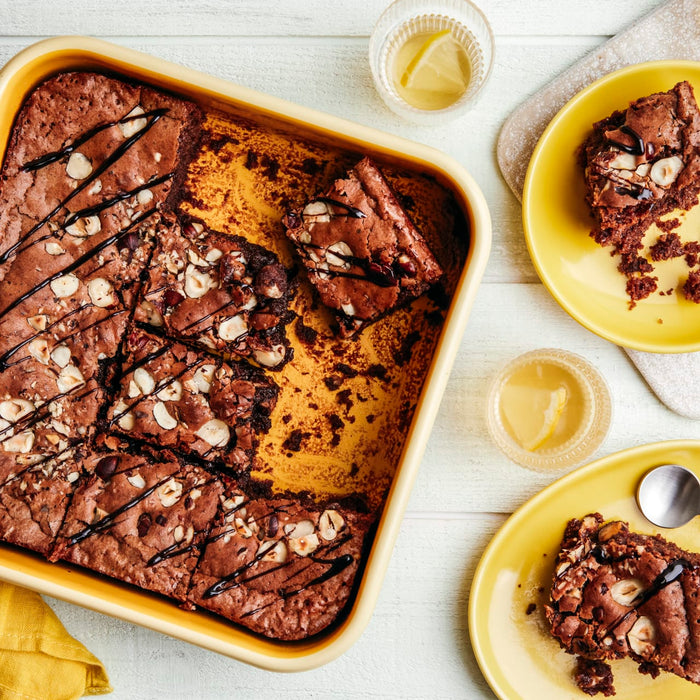 Un plat de brownies fraîchement cuits avec des noix et un filet de chocolat est présenté à côté de deux assiettes contenant des portions de brownies et des verres d'une boisson de couleur claire.