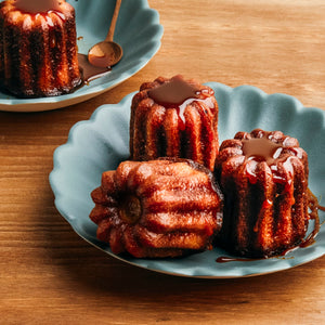 Trois cannelés sont disposés sur une assiette bleue avec un aspect brillant et caramélisé.