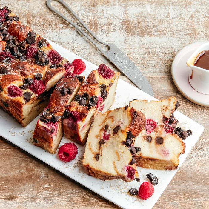 Une miche de pain perdu aux framboises et aux pépites de chocolat tranchée est posée sur un plat blanc avec des framboises fraîches et un petit ramequin de sauce.