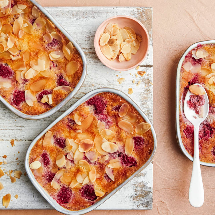 Trois petits gâteaux rectangulaires aux framboises et aux amandes sont présentés sur une planche en bois avec un bol d'amandes effilées.