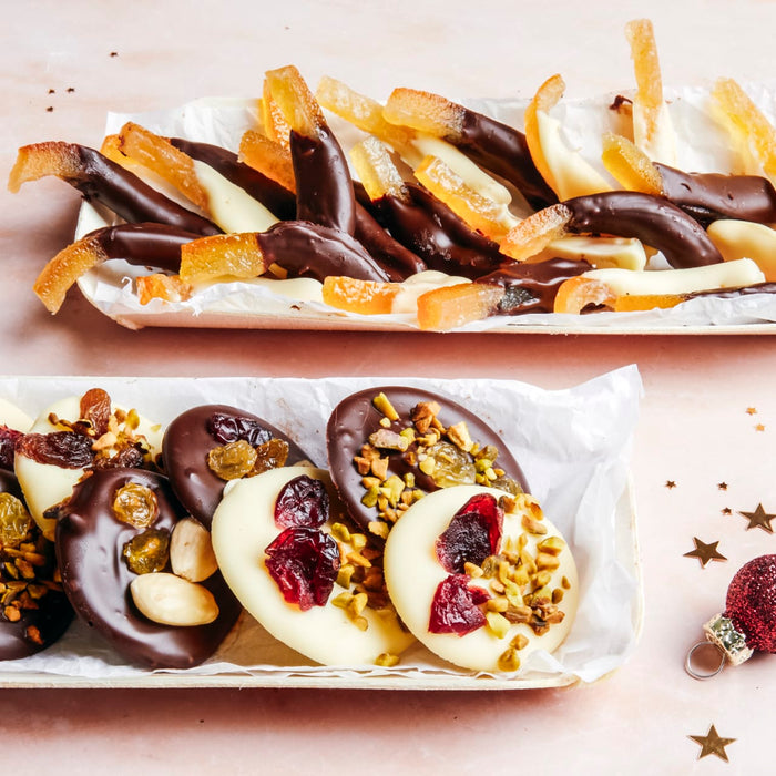 Deux plateaux présentent des écorces d'orange confites enrobées de chocolat et des ovales de chocolat garnis de fruits secs et de noix, mettant en valeur des friandises festives faites maison.