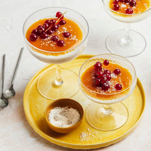 Un dessert visuellement attrayant mettant en vedette une panna cotta nappée d'une sauce orange vive et de groseilles rouges fraîches, servie dans d'élégants verres à pied.
