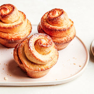 Trois viennoiseries dorées et tourbillonnées sont présentées sur une assiette rose clair, mettant en valeur leurs couches délicates et leur aspect invitant.