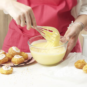 Une personne portant un tablier rose fouette une crème jaune dans un bol en verre, avec des pâtisseries à proximité, suggérant une scène de pâtisserie ou de cuisine.