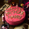 Un gâteau d'anniversaire sur le thème de Harry Potter avec un glaçage rose et des lettres vertes qui disent Happee Birthday Harry, entouré de bougies et de décorations sur le thème de Harry Potter.