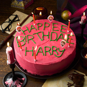 Un gâteau d'anniversaire sur le thème de Harry Potter avec un glaçage rose et des lettres vertes qui disent Happee Birthday Harry, entouré de bougies et de décorations sur le thème de Harry Potter.